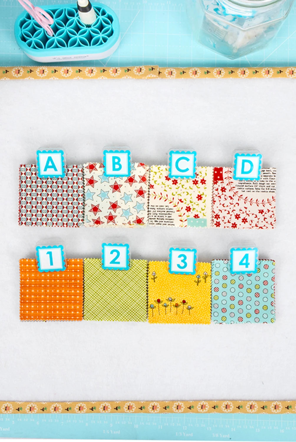 Colorful fabric swatches labeled A to D on a light blue background with sewing Quilt Clips with letter tiles and fabric on a blue surface tools.