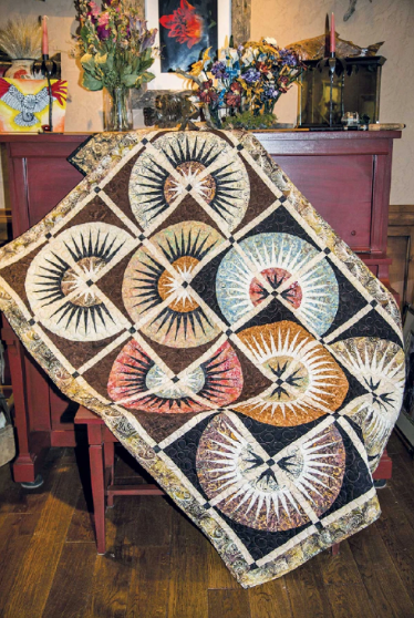 Quilt with geometric pattern draped over a chair in a room with floral arrangements on a table. Clockworx cut Loose Quilt Pattern - By Judy Niemeyer - Carmon's Quilt Shop
