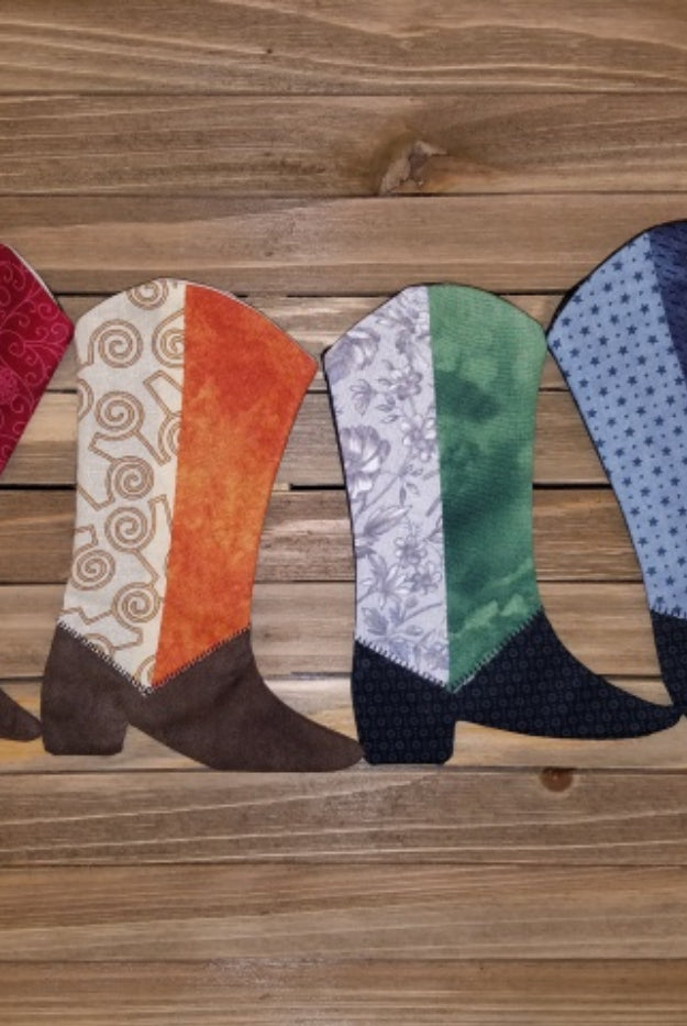 A series of quilted fabric boots in various colors arranged on a wooden Just Kickin' It Quilt Pattern - Moderns Quilt Pattern by Glenda Wilder - Carmon's Quilt Shopsurface.