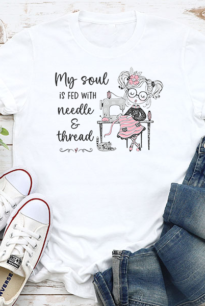 White t-shirt with sewing-themed design and text, paired with jeans and sneakers on a wooden surface.