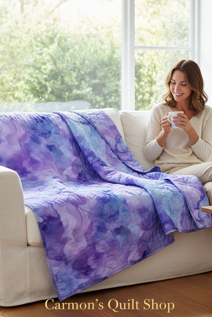 Carmon's Quilt Shop Aura Watercolor purple cotton quilt draped over a white sofa with a woman holding a mug.
