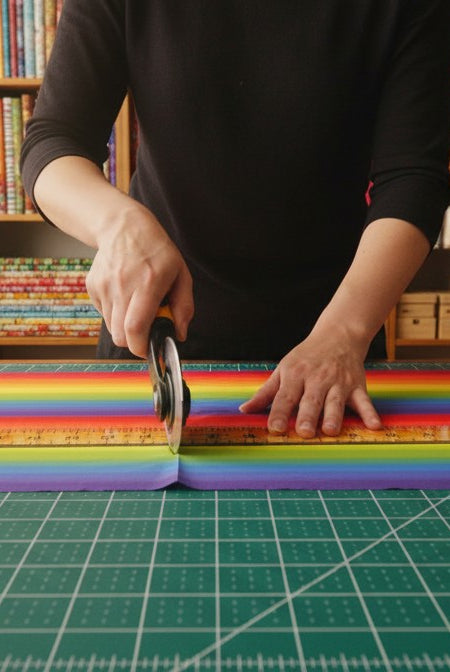 Carmon's Quilt Shop rainbow ombre cotton fabric being cut with a rotary cutter on a green cutting mat in a studio.