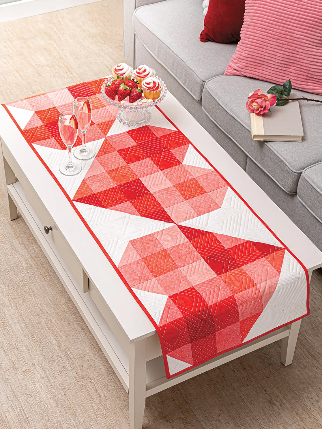 Carmon's Quilt Shop red and white geometric quilted table runner on a coffee table with strawberries and cupcakes.