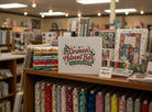 Carmon's Quilt Shop limited-edition advent box displayed on a wooden shelf with stacks of colorful quilting fabrics.