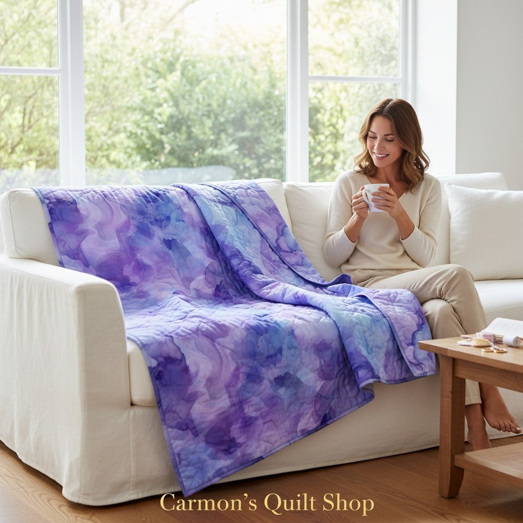 Carmon's Quilt Shop Aura Watercolor purple cotton quilt draped over a white sofa with a woman holding a mug.