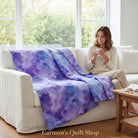 Carmon's Quilt Shop Aura Watercolor purple cotton quilt draped over a white sofa with a woman holding a mug.