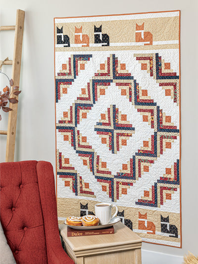 Carmon's Quilt Shop autumn wall quilt featuring geometric patchwork and cat motifs hanging above a cozy armchair.