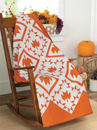 Carmon's Quilt Shop autumn quilt with orange and white maple leaf patchwork draped over a wooden rocking chair.