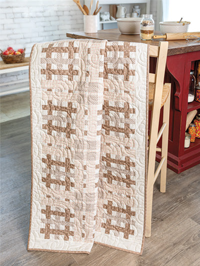 Carmon's Quilt Shop autumn quilt featuring brown and cream patchwork patterns draped over a wooden chair in a kitchen.