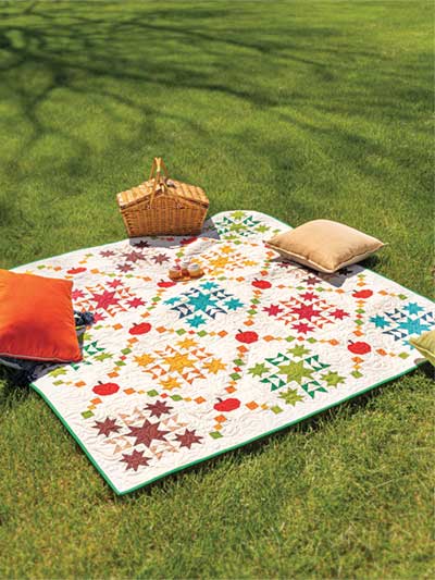 Carmon's Quilt Shop autumn quilt featuring colorful patchwork stars and apple motifs on a grassy lawn with a picnic basket