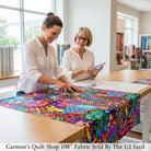 Two women at Carmon's Quilt Shop cutting vibrant QT Fabrics Carnivale floral geometric cotton fabric on a table.