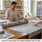 Carmon's Quilt Shop staff cutting grey mottled Panorama Rapture blender cotton fabric from a wide bolt in a studio.