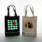 Carmon's Quilt Shop Halloween tote bags, one black with glow-in-the-dark pumpkins, one cream with a black cat and pumpkins.