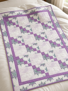 Carmon's Quilt Shop Dragonfly Wonder lavender quilt featuring geometric blocks and floral prints on a white bed.