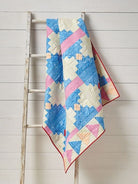 Carmon's Quilt Shop Log Cabin Variations quilt featuring blue, pink, and cream geometric patterns on a wooden ladder.