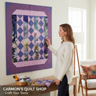 Woman admiring a floral quilt wall hanging from Carmon's Quilt Shop with purple and lavender diamond patterns.