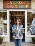 Woman reading the 2026 All Missouri Shop Hop Magazine outside Carmon's Quilt Shop with quilt patterns visible.