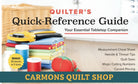 Carmon's Quilt Shop Quilter's Quick-Reference Guide with rotary cutter, fabric stack, and pincushion on a desk.