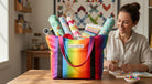 Carmon's Quilt Shop rainbow ombre tote bag filled with fabric rolls on a wooden table with a woman crafting nearby.