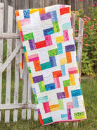 Carmon's Quilt Shop Scrap-Happy quilt pattern featuring a colorful geometric design draped over a white garden fence.
