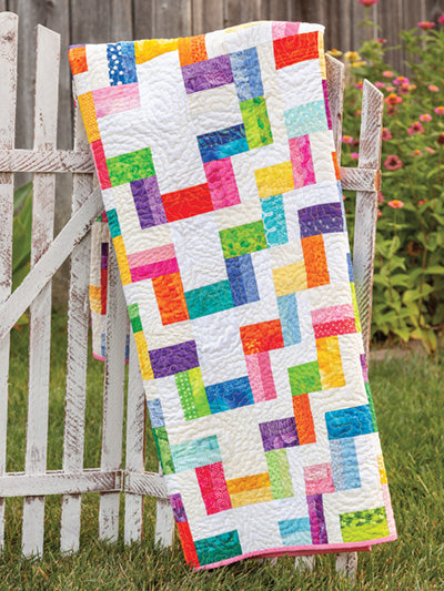 Carmon's Quilt Shop Scrap-Happy quilt pattern featuring a colorful geometric design draped over a white garden fence.